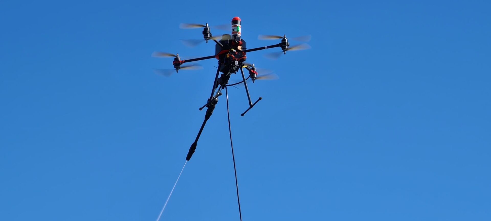 Drone relié au sol par un tuyau, projetant un jet fin par sa lance, sur fond de ciel bleu